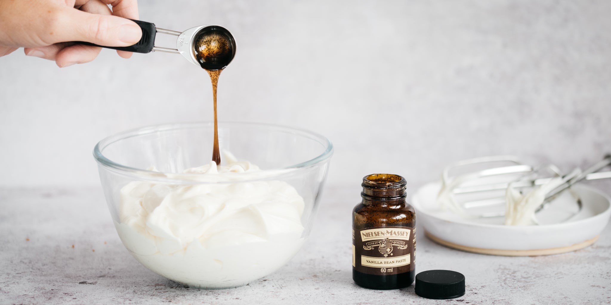 Glass bowl of buttercream icing and vanilla bean paste being poured in with a measuring spoon. Jar of vanilla next to the bowl