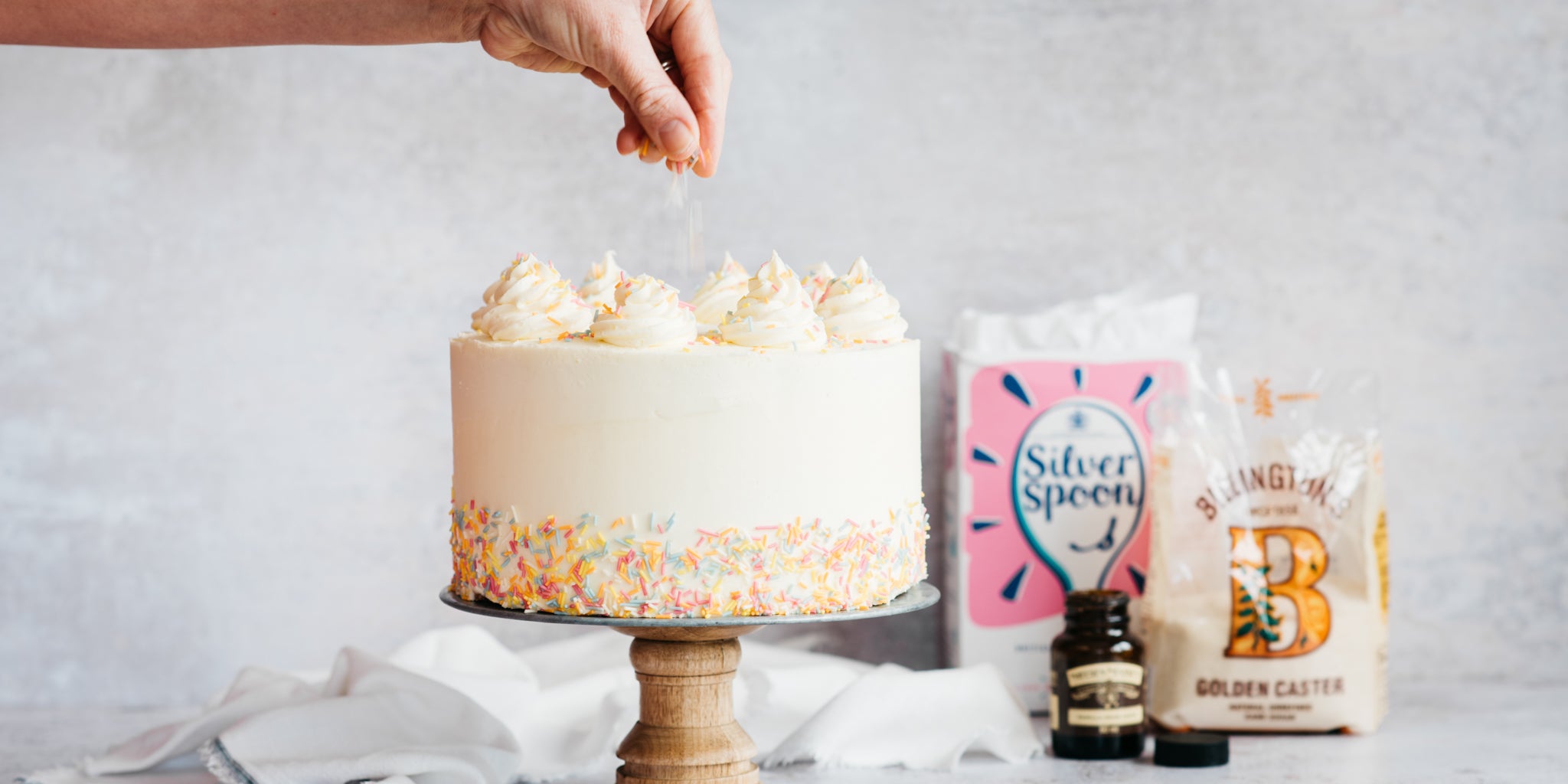 Vegan Vanilla Cake being hand sprinkled with decoration, with a bag of Silver Spoon Icing Sugar and Caster sugar in the background