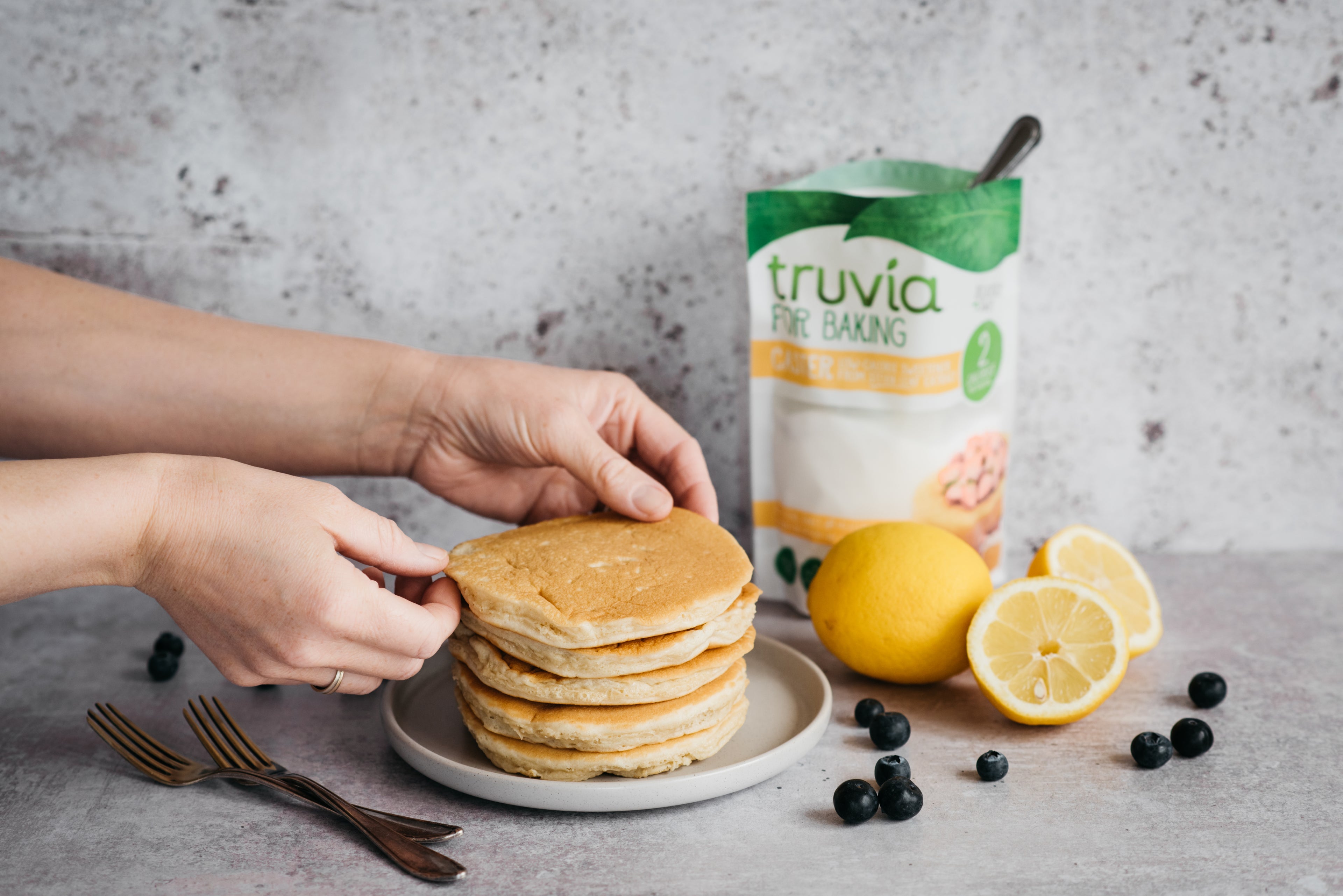 Hands stacking cinnamon pancakes, next to scattered blueberries and a bag of Truvia for Baking Caster