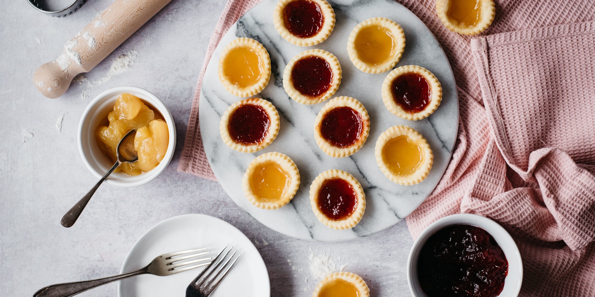 Top view of Apricot & Strawberry Jam Tarts served on a marble board, next to a bowl of jam and a rolling pin