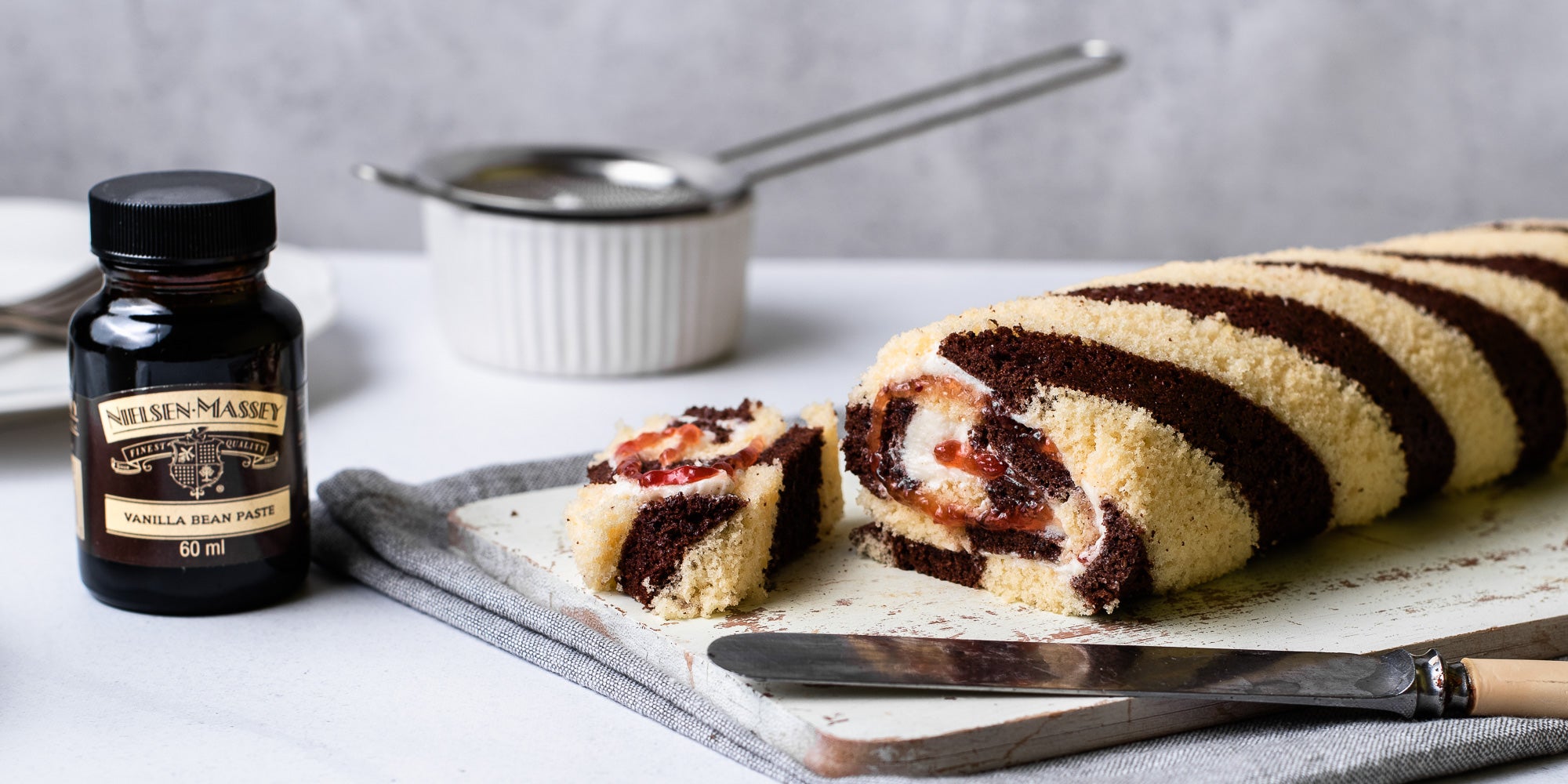 A slice of Chocolate & Vanilla Swiss Roll served on a wooden board, next to a bottle of Nielsen-Massey Vanilla Extract and a ramekin holding a sieve in the background