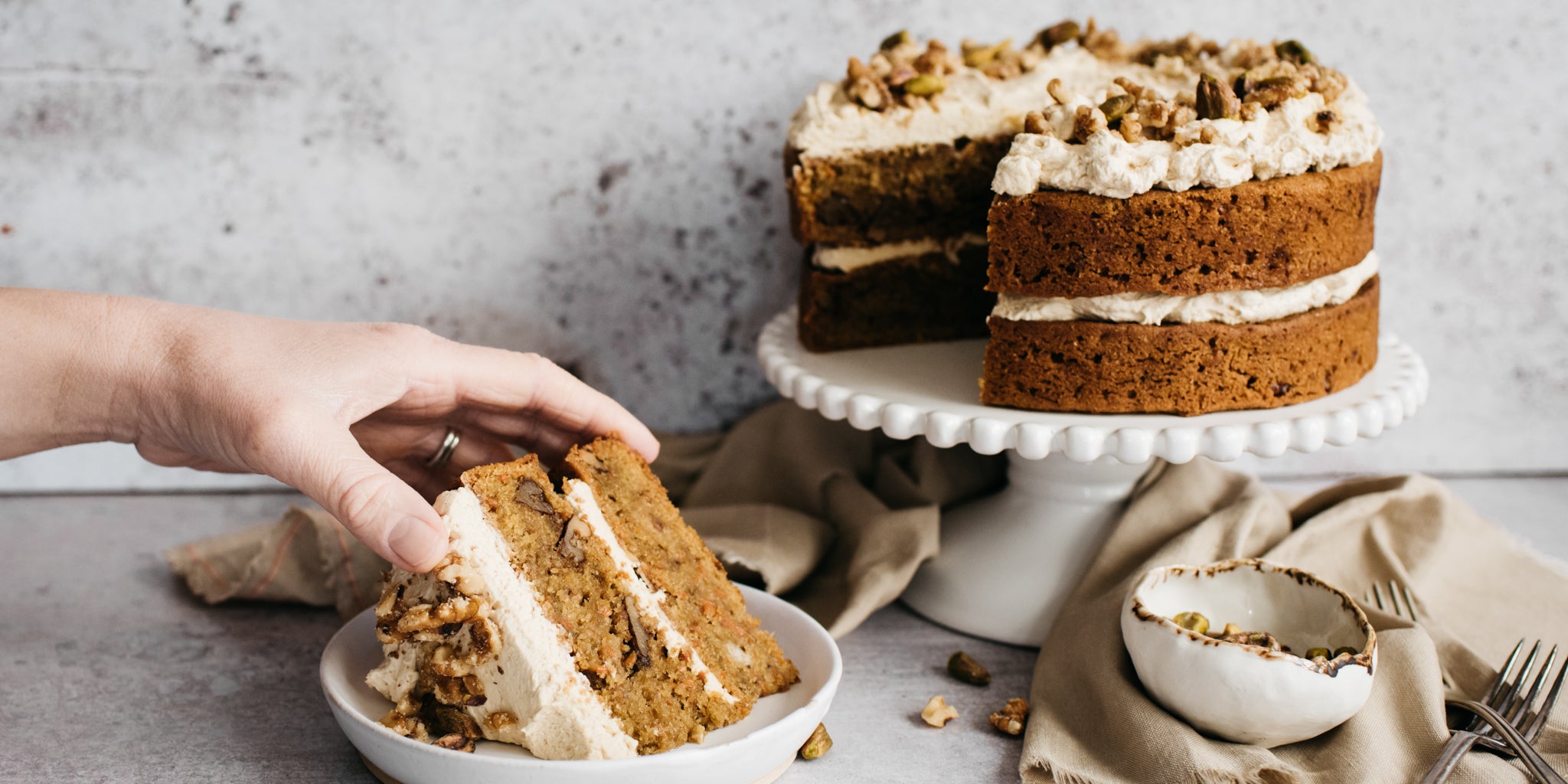 Vegan Carrot Cake with a slice cut out of it, served on a plate with a hand holding the Vegan Carrot Cake ready to bite into