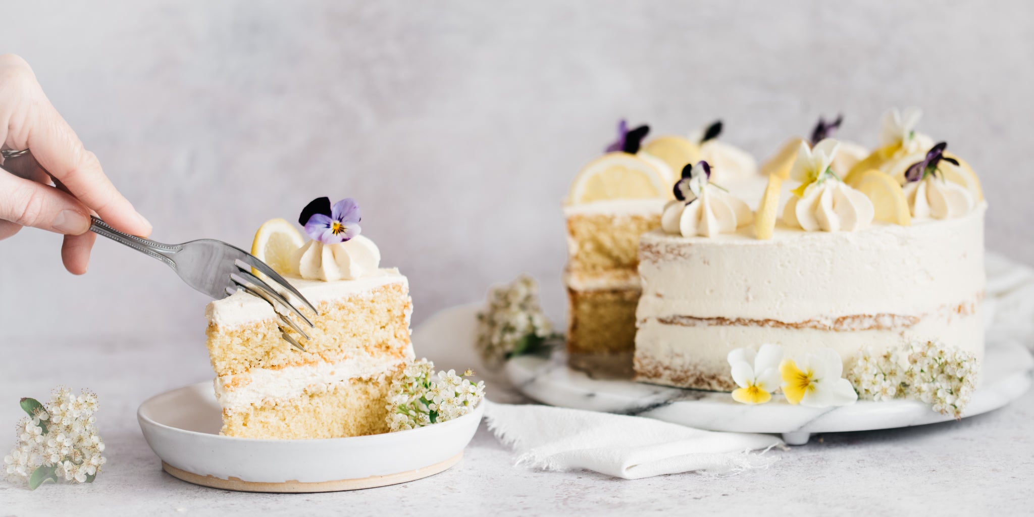 Sponge cake decorated with flowers with a slice removed and on a white plate in the foreground. Hand reaching in to take a bite of the slice