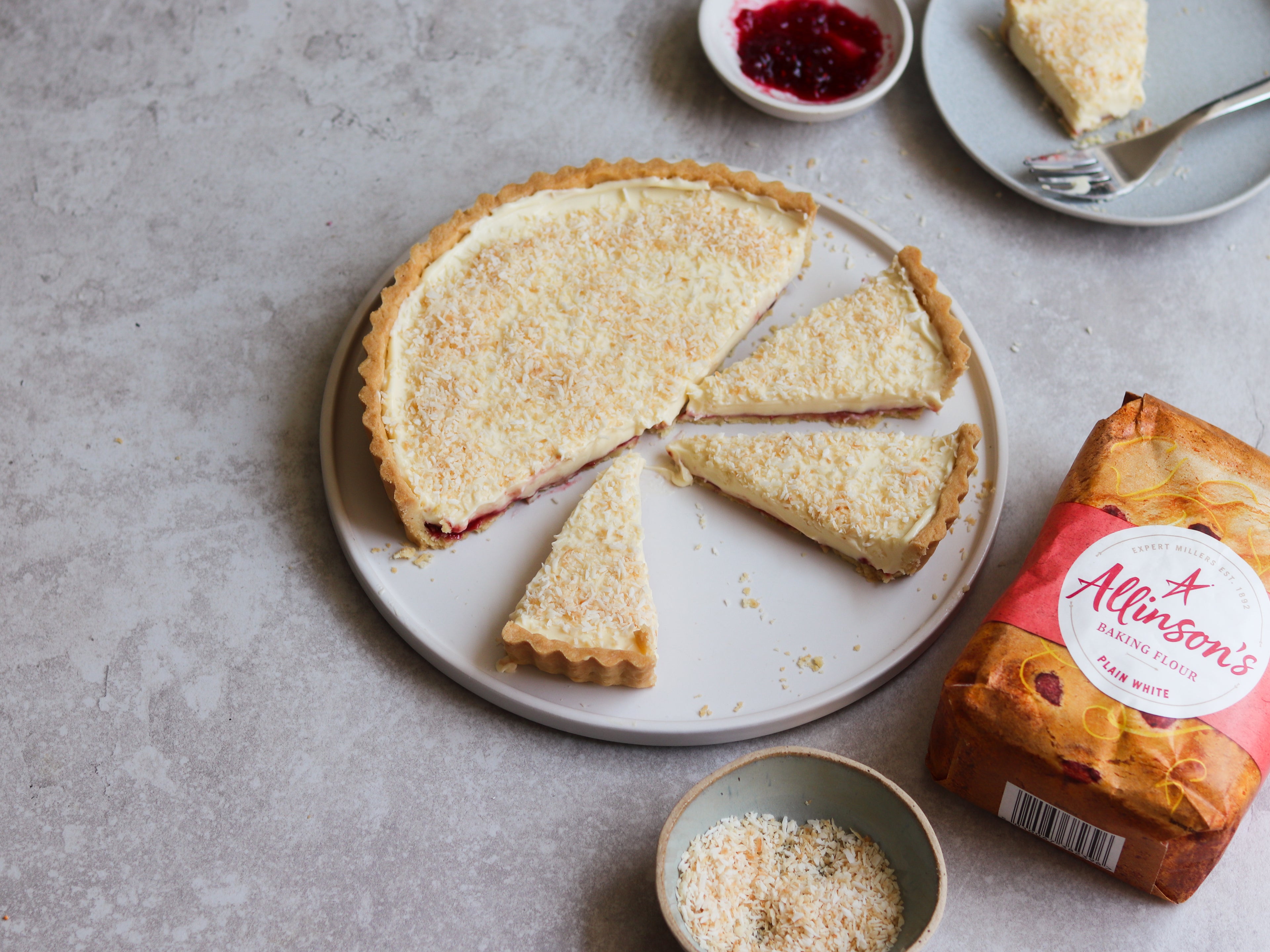 Manchester Tart sliced into servings, on a plate. Next to a bowl of jam and toasted coconut, and a bag of Allinson's Plain Flour