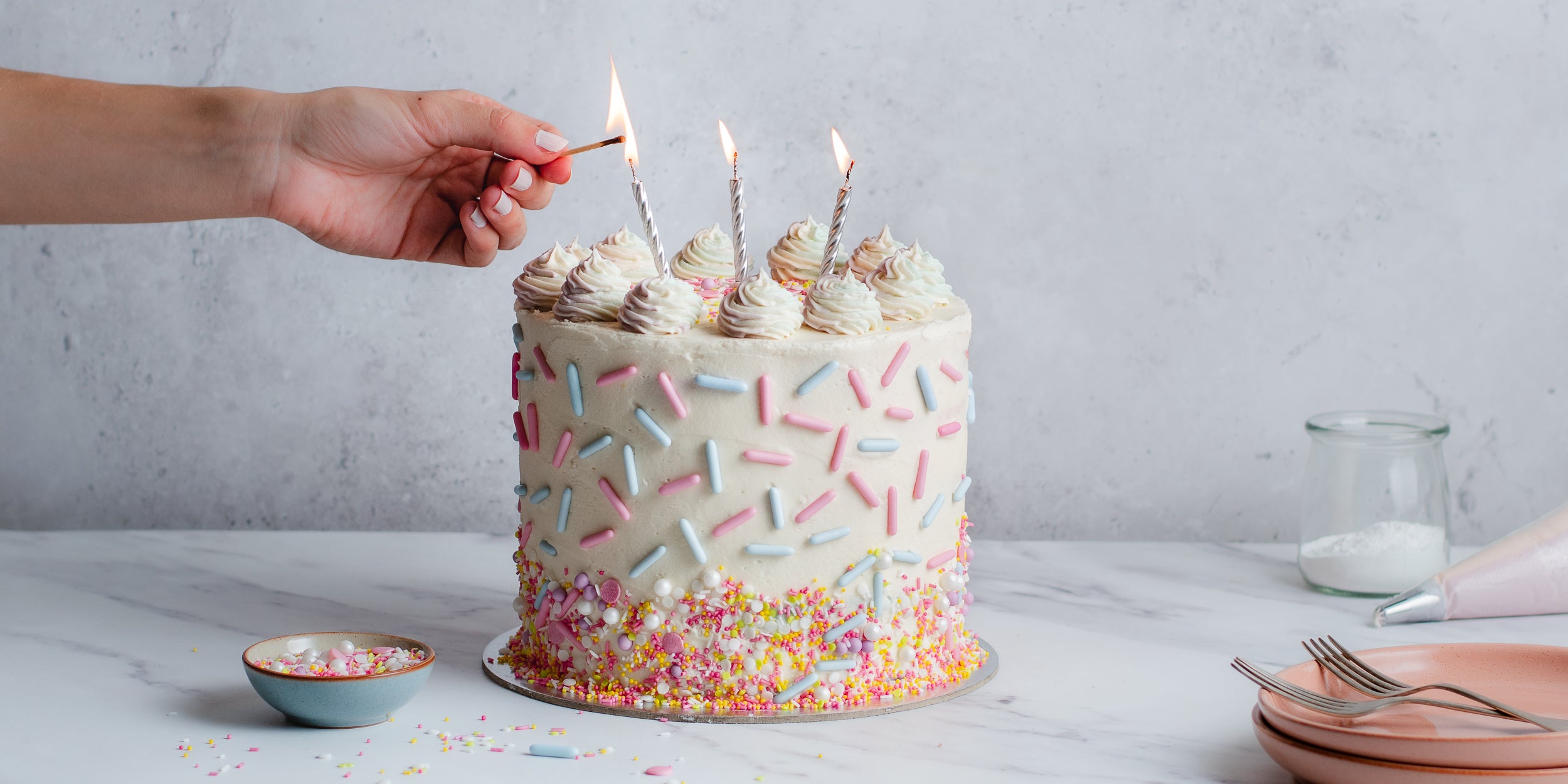Hand reaching in with a match to light candles on top of white sprinkle covered birthday cake