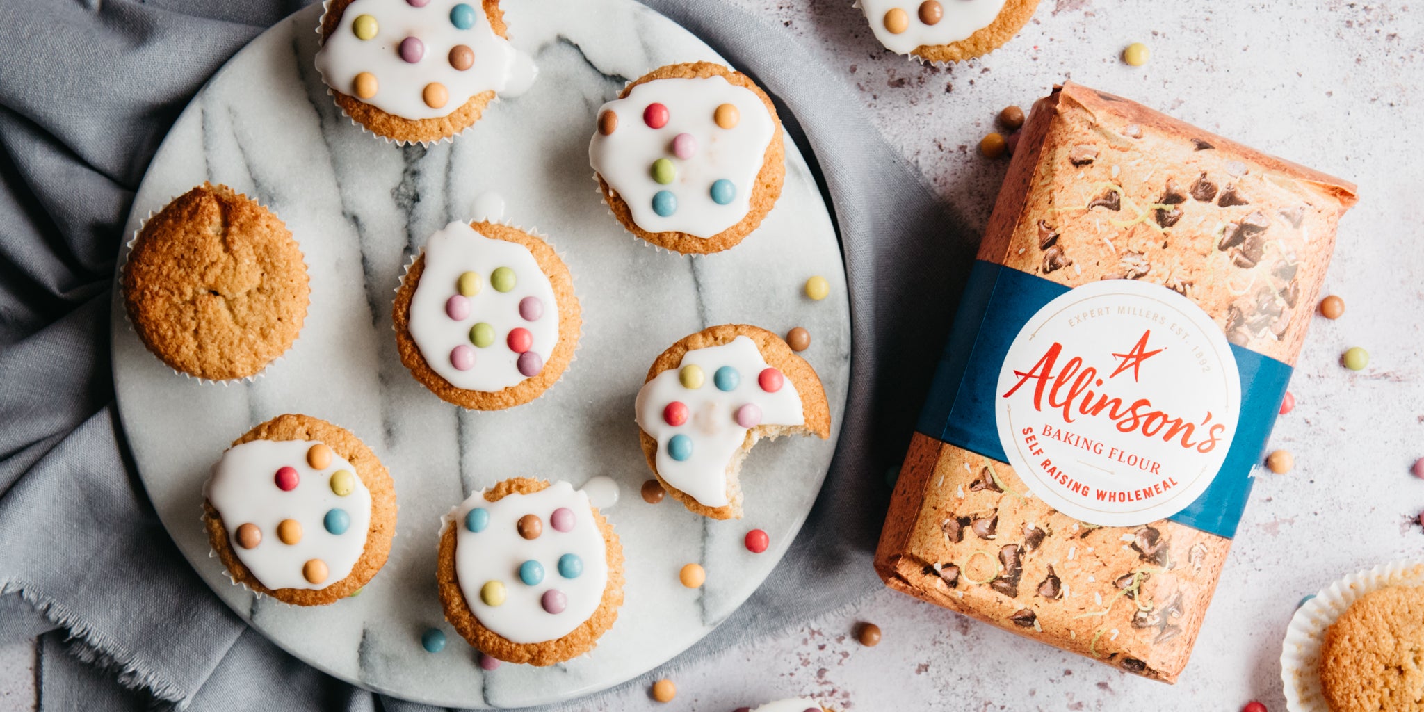 Wholemeal Fairy Cakes on a marble cake stand, next to a bag of Allinson's Wholemeal flour, and a cupcake with a bite taken out of it.