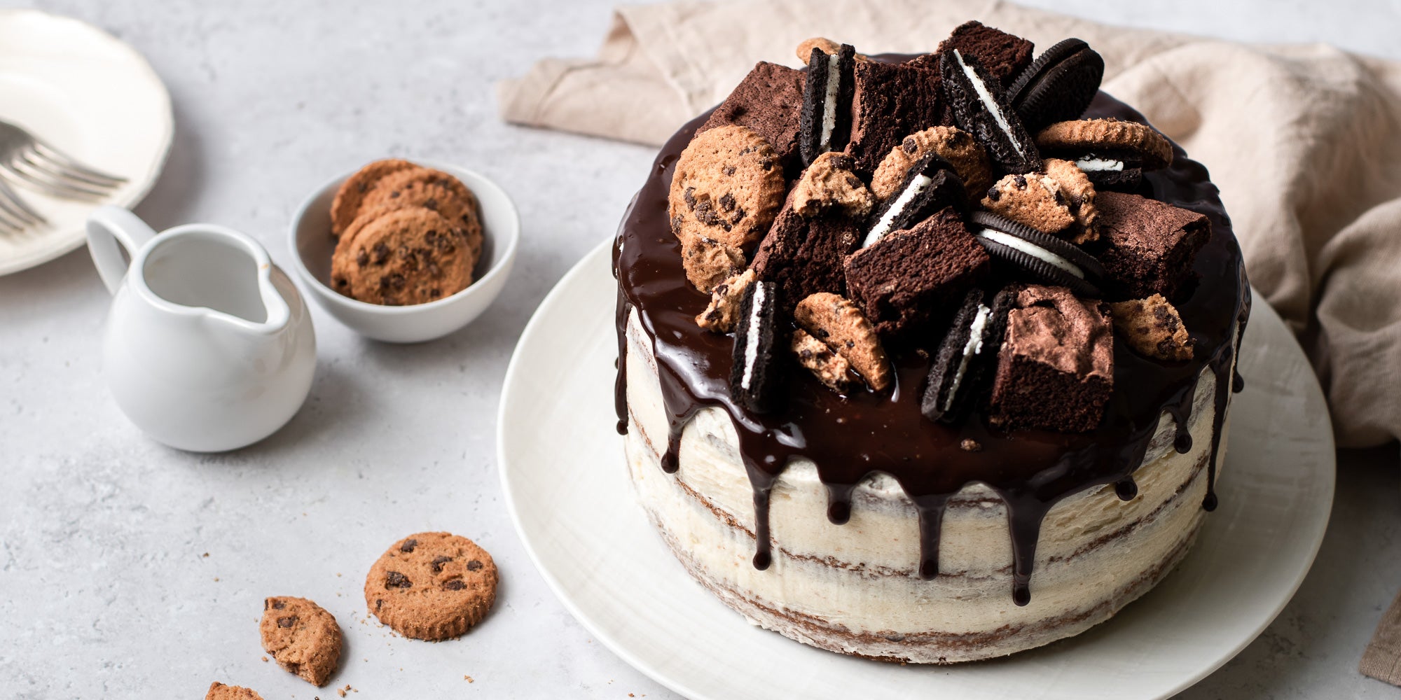Cookie, Oreo & Brownie Layered Cake topped with oreos and cookies, next to a bowl of mini cookies and a small jug of chocolate ganache