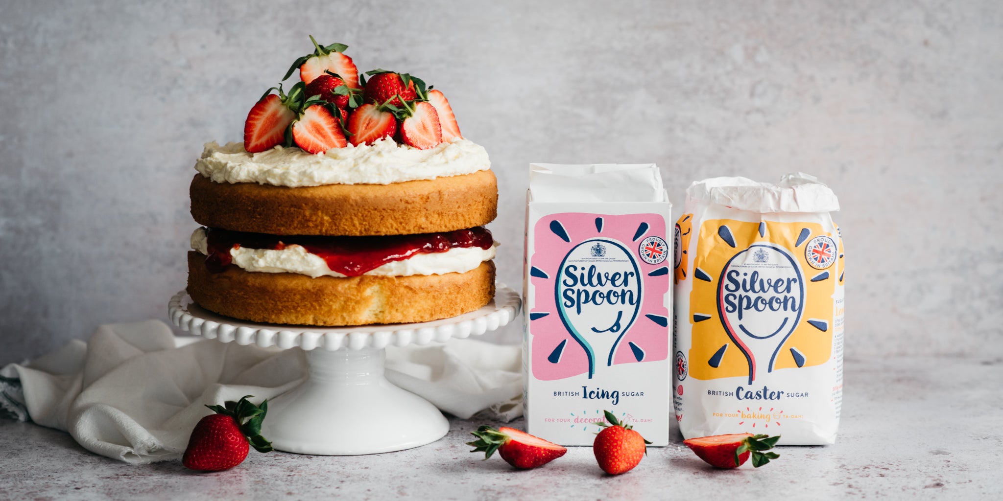 Show-stopping Gluten Free Victoria Sponge served on a cake stand and topped with sliced strawberries. Next to a bag of Silver Spoon Icing Sugar and Silver Spoon Caster Sugar