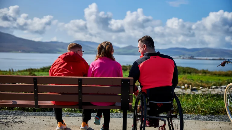 People sitting on a bench and one person in a wheelchair with coastal view