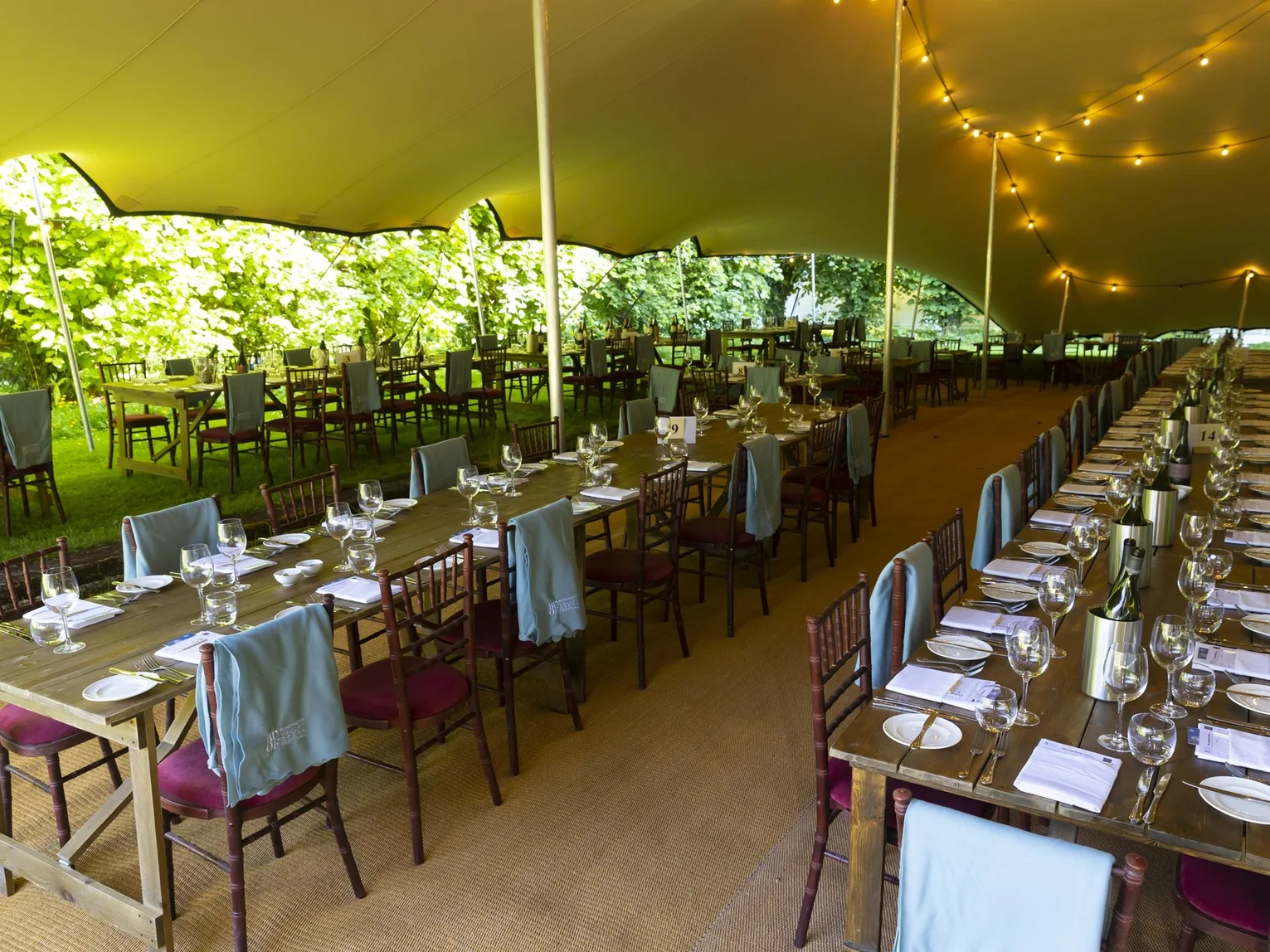 Interior of a large outdoor marquee set for a dinner event with long wooden tables, wine glasses and festoon lighting