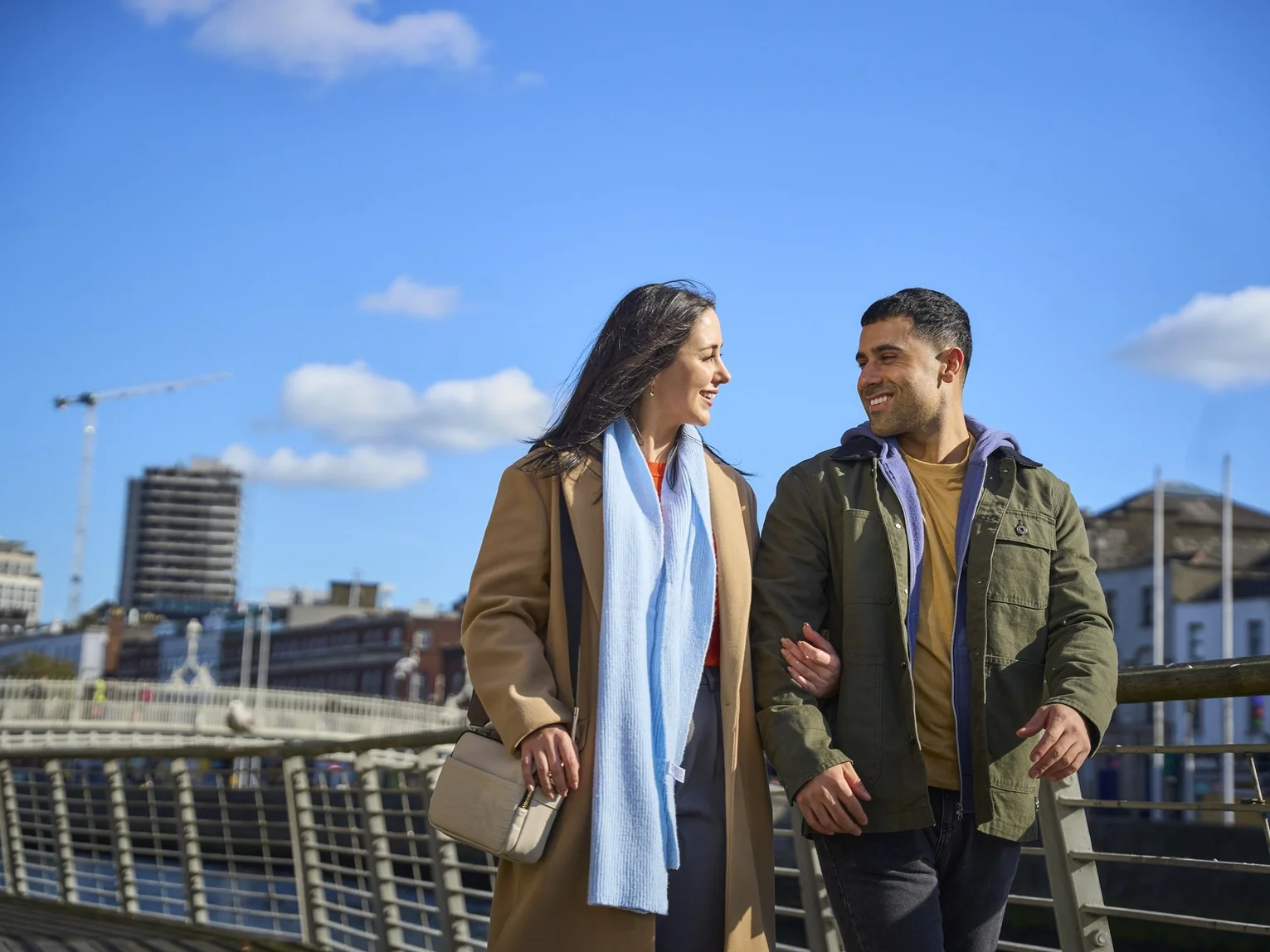 A couple walking arm-in-arm across a bridge in Dublin city centre on a bright sunny day with a blue sky.