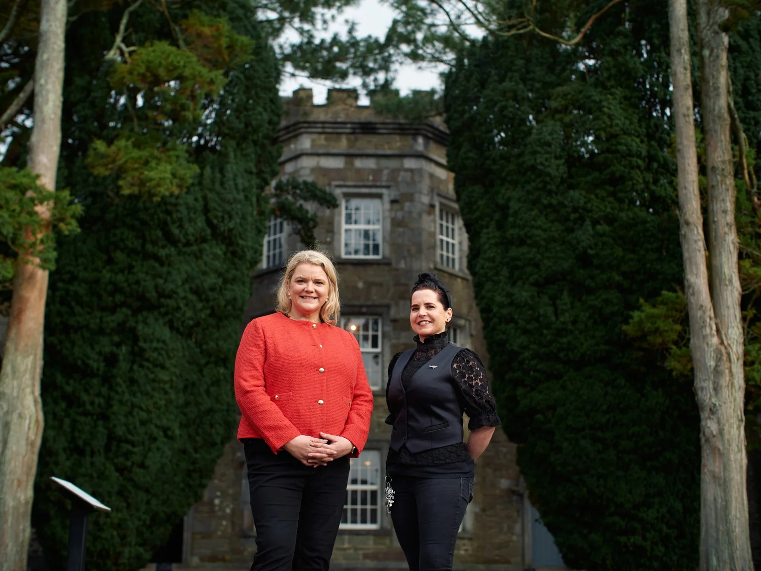 Two people standing outdoors in front of a historic stone manor house surrounded by tall trees