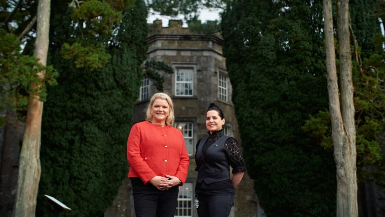 Two people standing outdoors in front of a historic stone manor house surrounded by tall trees