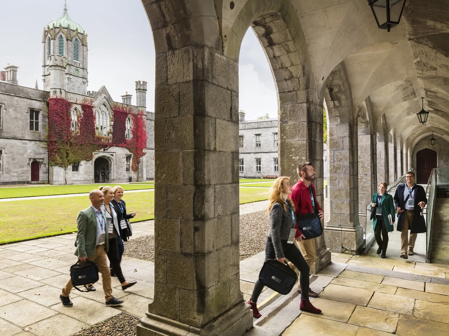 Tourism professionals walking through the stone cloisters of a historic building with red ivy-clad walls in the background.