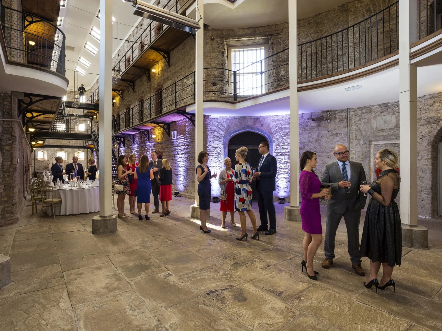 Guests networking at a formal drinks reception in a historic stone-walled venue Cork Jail with iron balconies and atmospheric lighting
