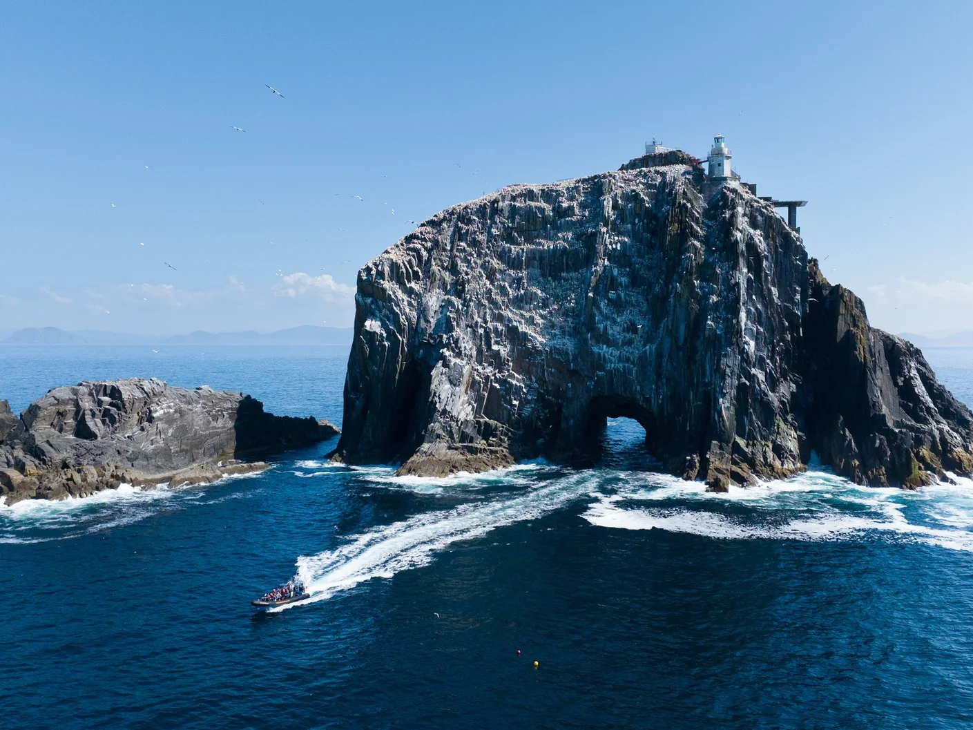 Aerial view of a dramatic sea stack rock island with a lighthouse, surrounded by crashing waves and a tourist boat