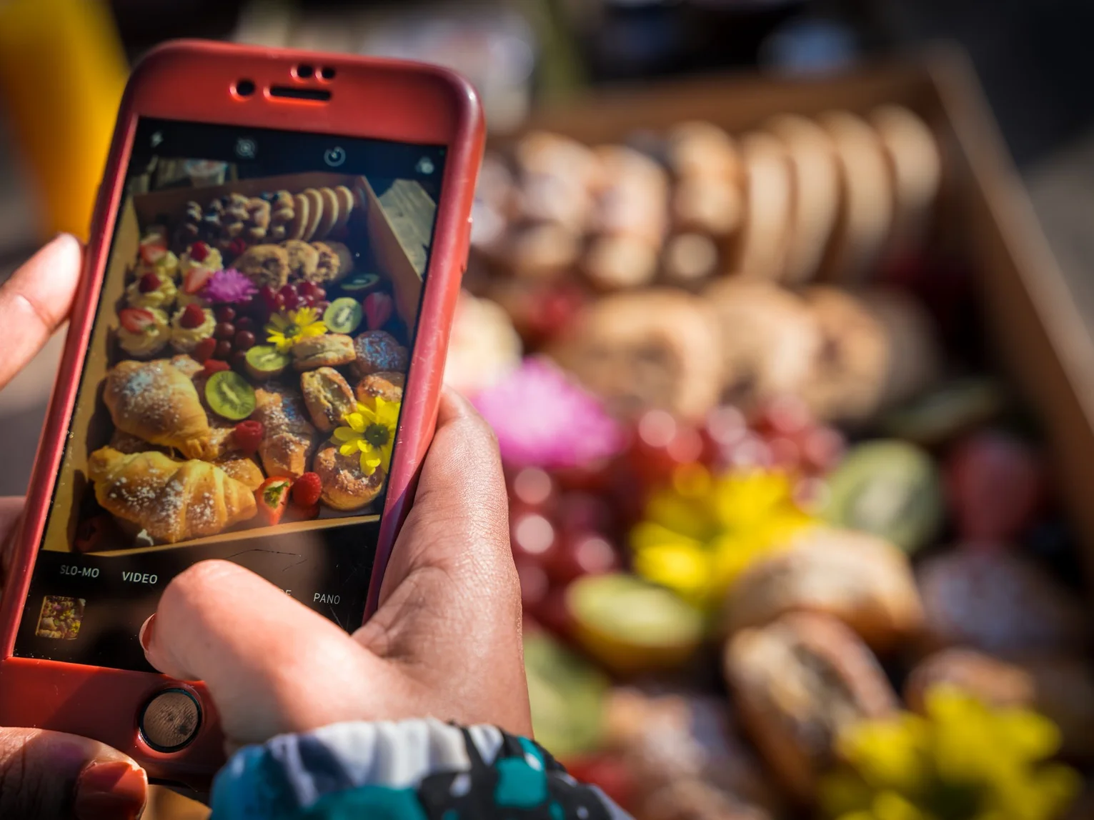 Person photographing a colourful arrangement of pastries and fruit at an outdoor market using a smartphone