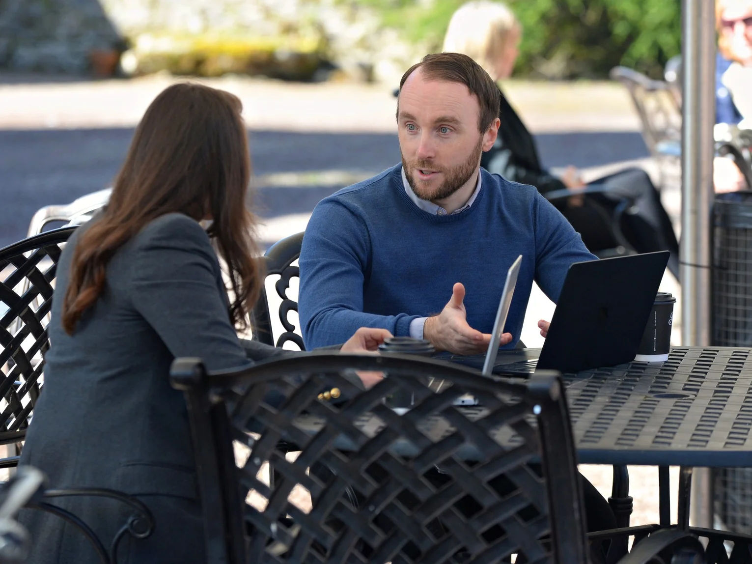 Two people having an outdoor meeting at a table with a laptop and takeaway coffee