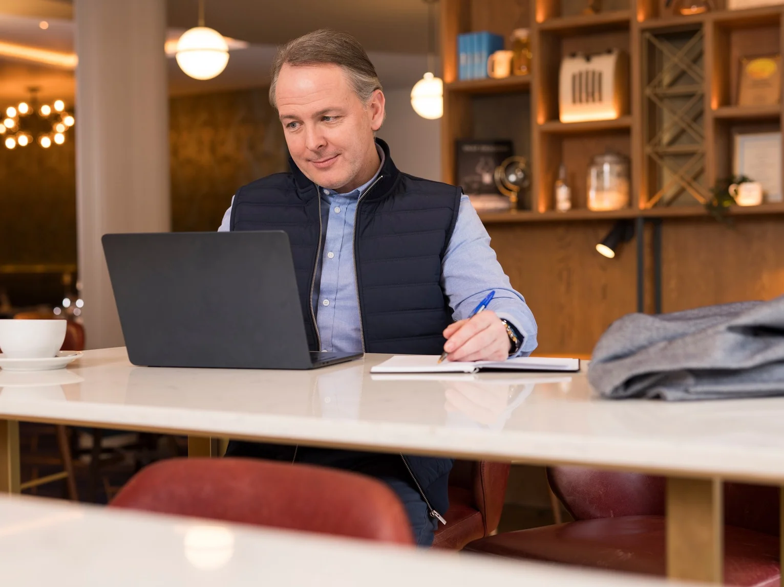Man in a navy gilet working at a laptop and taking notes in a hotel lounge