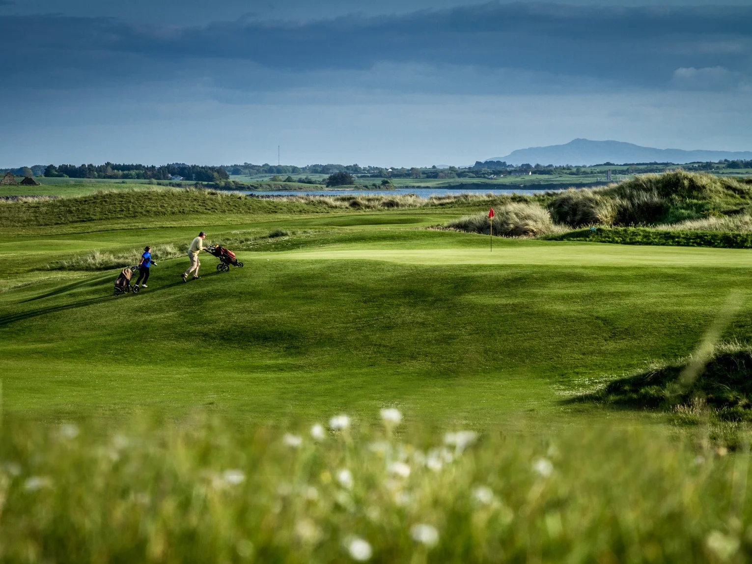 Two golfers pushing trolleys along a links fairway with a coastal lake and mountains in the distance
