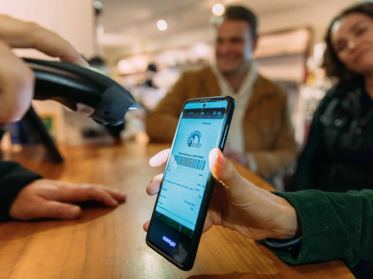 Person scanning a digital booking confirmation on a smartphone at a venue counter, with customers in the background