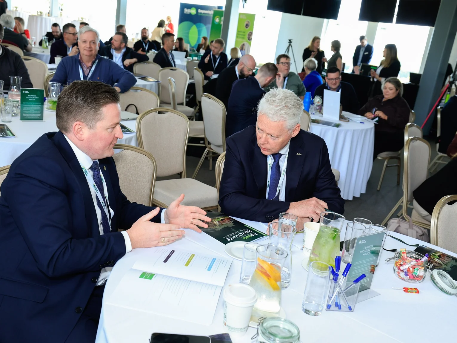 Two men in suits having a one-to-one business conversation at a conference table with other attendees in the background