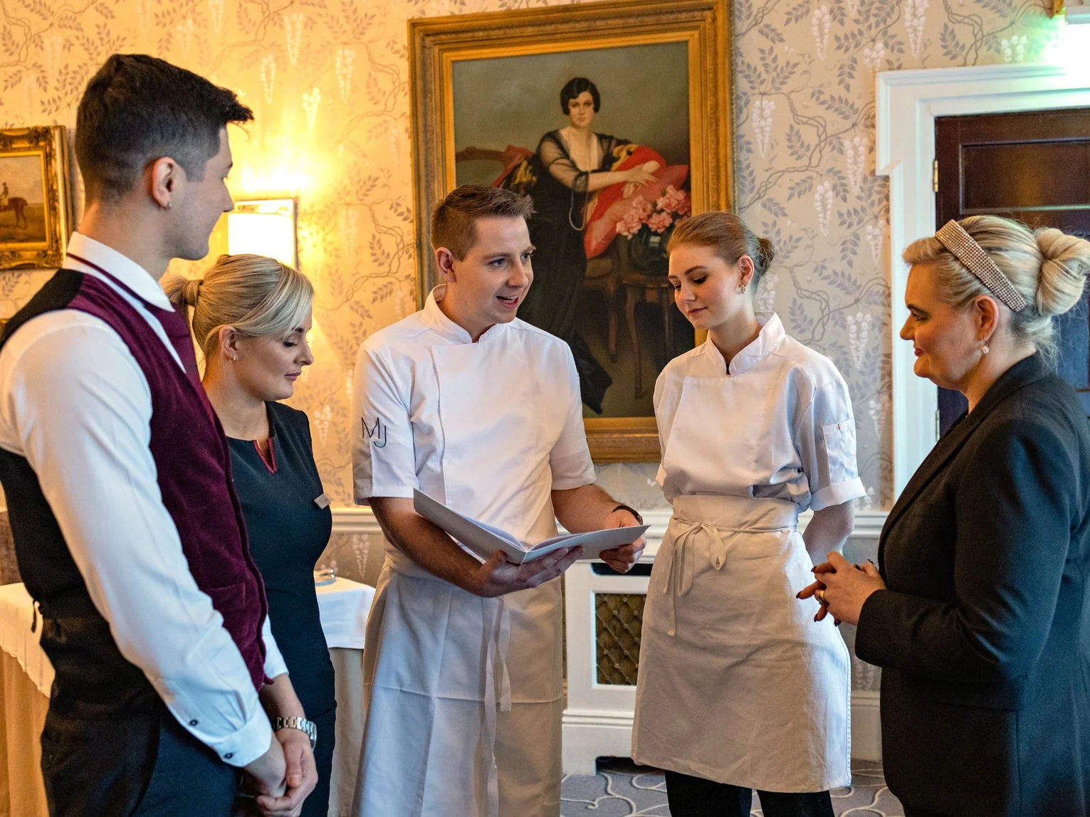 Five hospitality staff members including a chef in whites gathered in discussion in an elegantly decorated hotel room