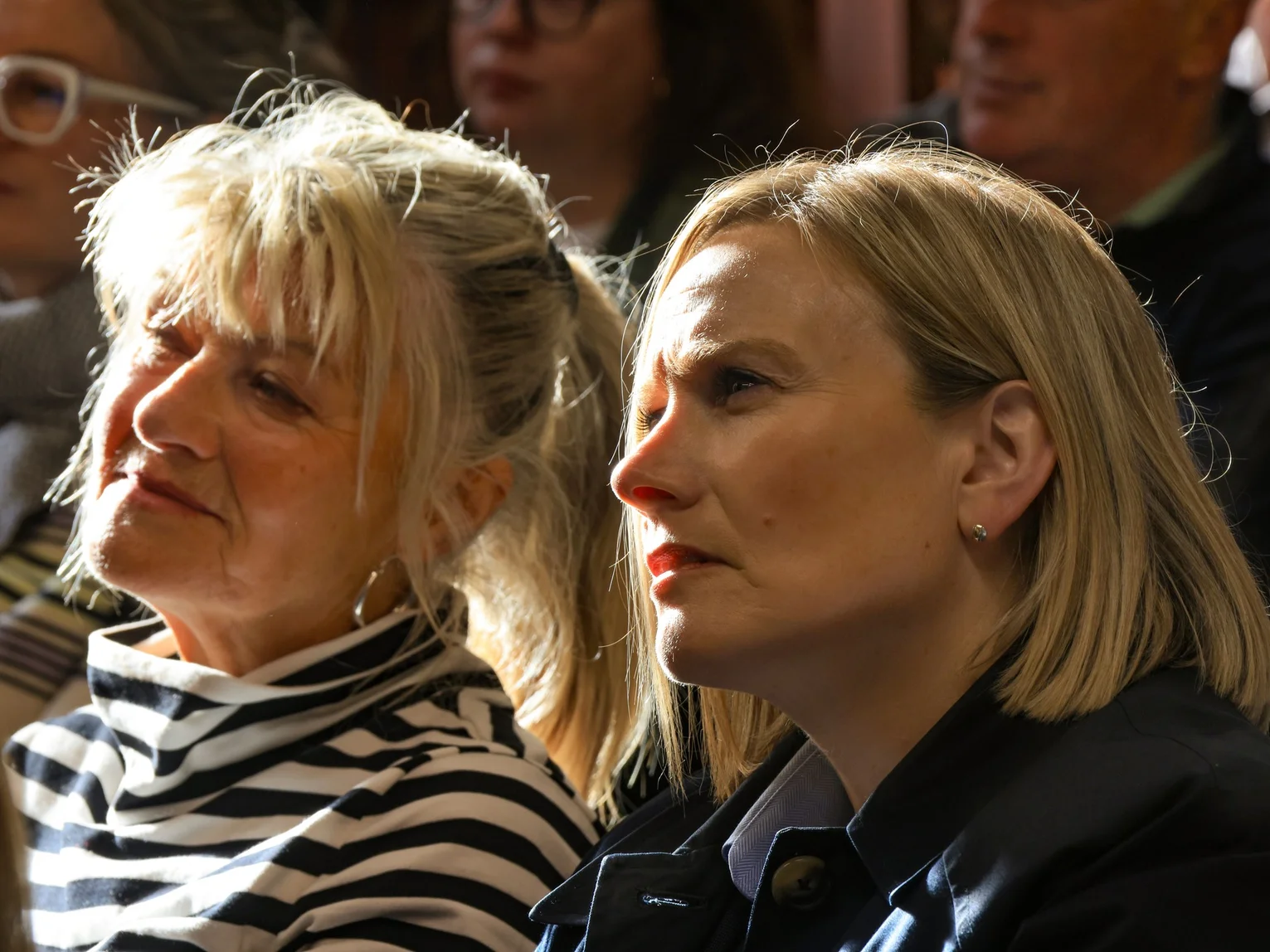 Two women seated in a sunlit audience listening attentively at a conference or event