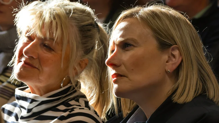 Two women seated in a sunlit audience listening attentively at a conference or event