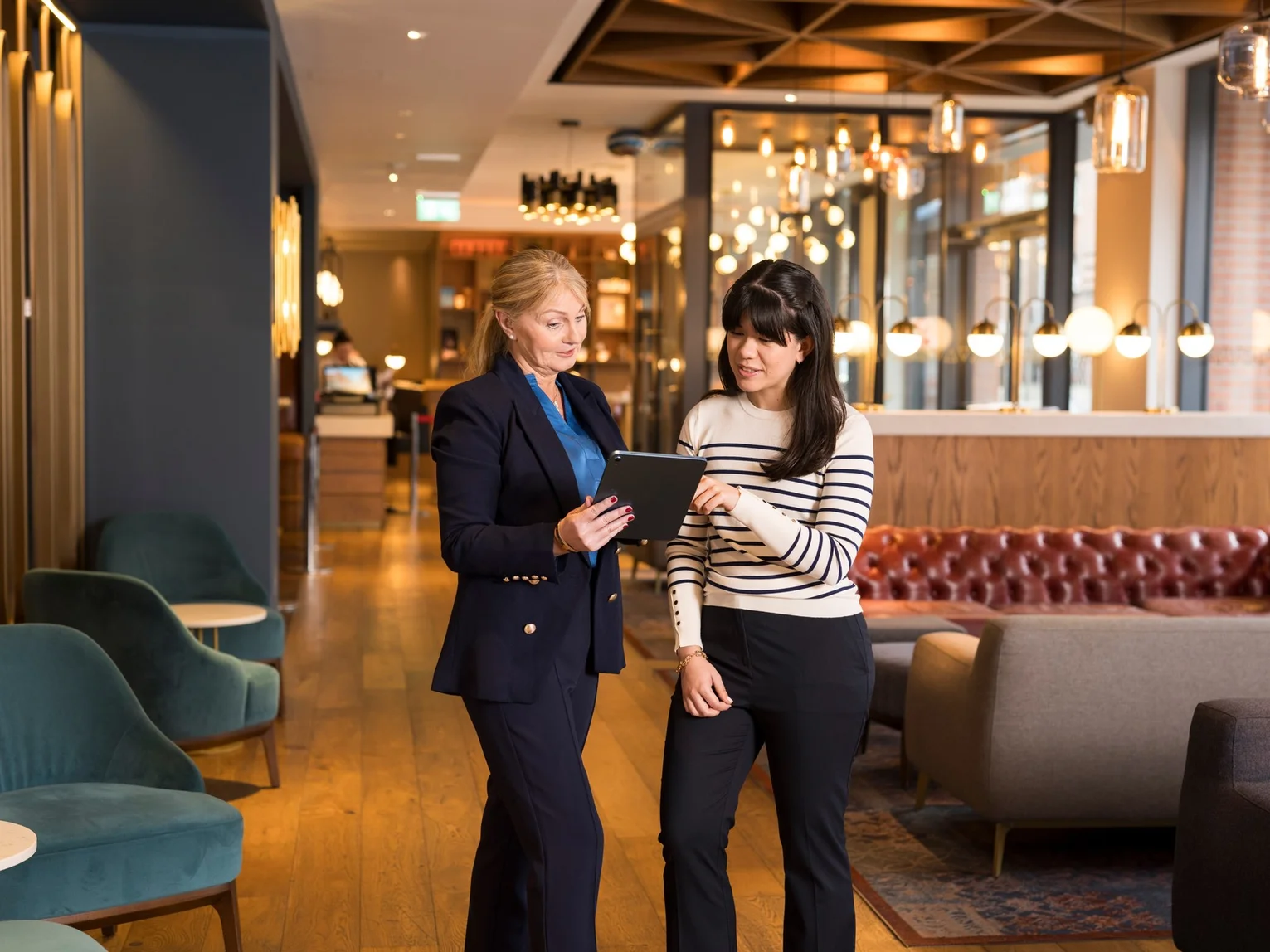 Two women reviewing content together on a tablet while standing in a hotel lobby lounge