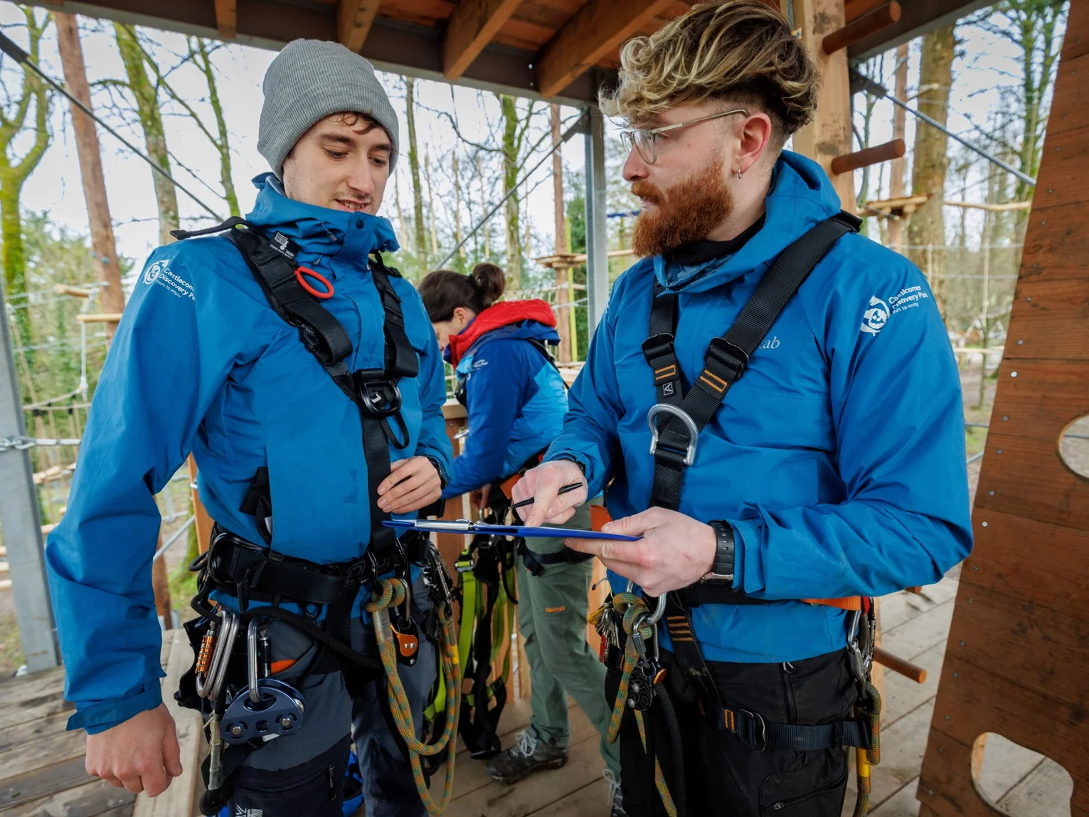 Two outdoor activity staff in harnesses checking safety equipment at a high ropes platform
