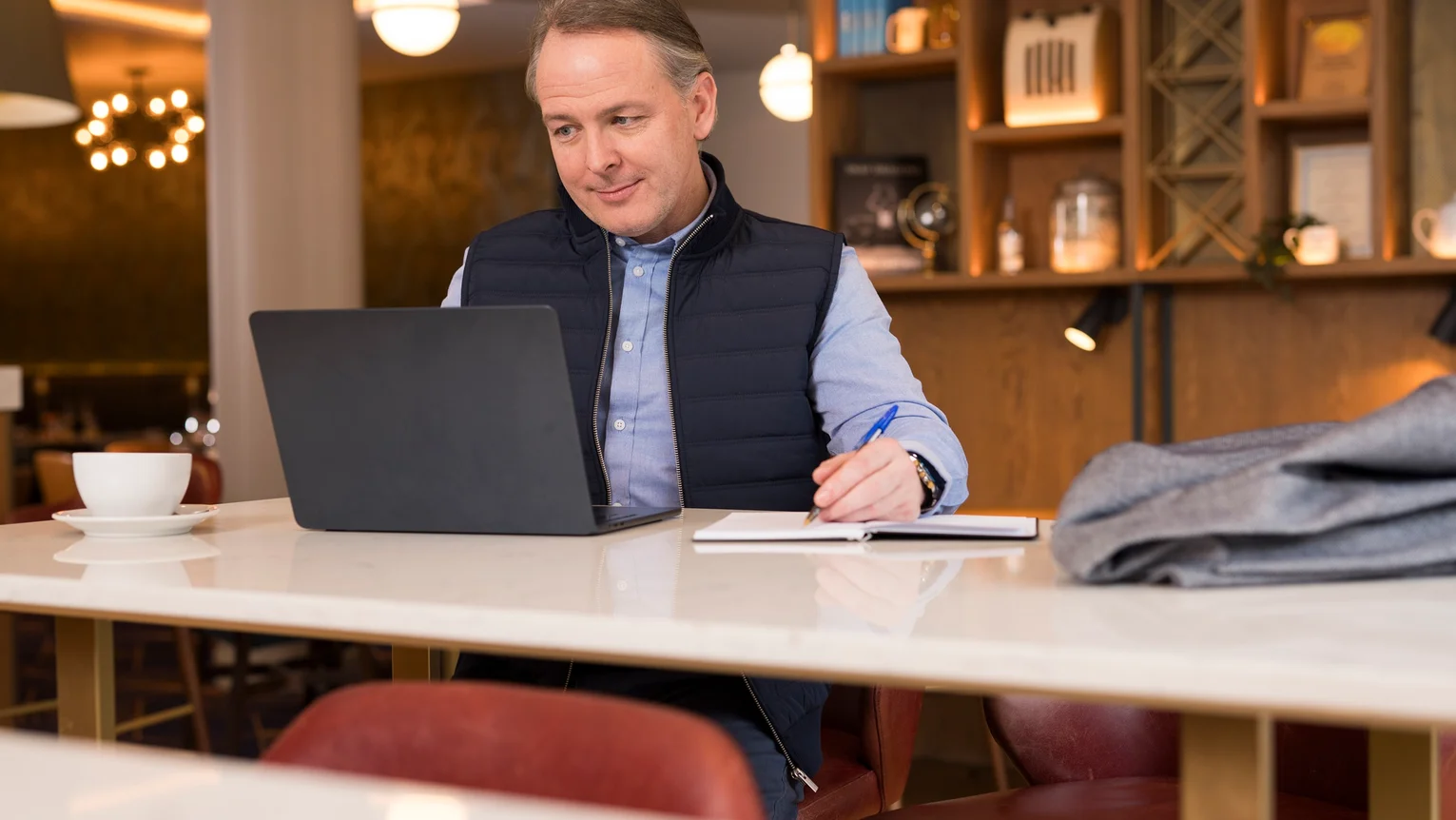 Smartly dressed man working on a laptop, in a hotel lobby, with a coffee.