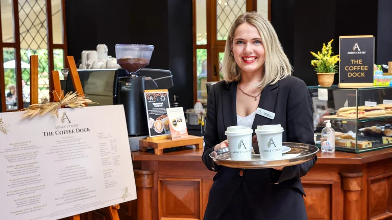 Smiling hotel staff member holding a tray of branded takeaway coffees at a café counter with menu board