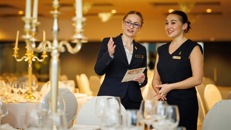 Two hotel staff members in formal uniforms speaking together in a set banquet dining room