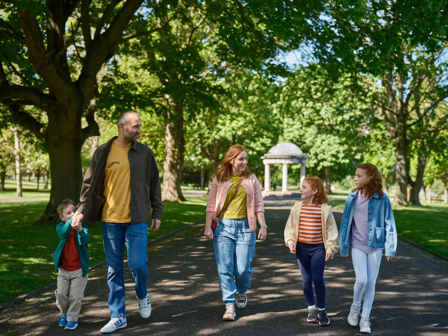 Family of five walking together along a tree-lined path in a public park on a sunny day