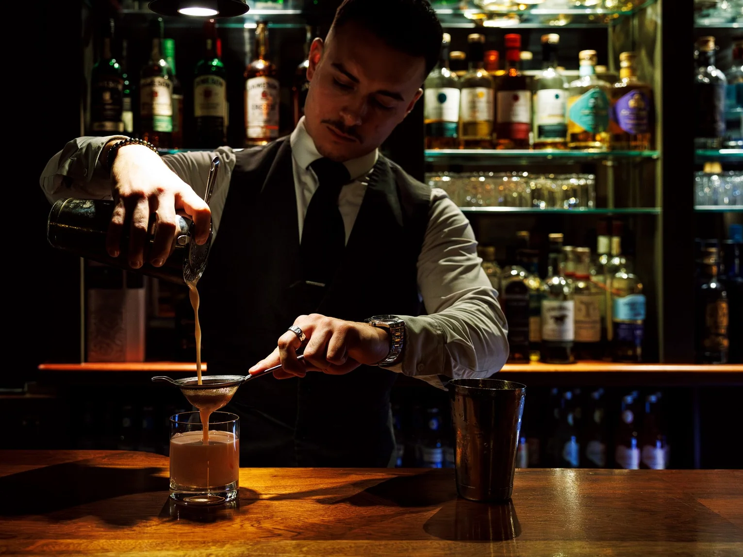 Bartender straining a cocktail into a glass at a moody, wood-panelled bar with backlit spirits shelves