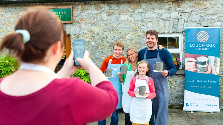 Person using a smartphone to photograph two attendees posing in front of a Fáilte Ireland branded event backdrop
