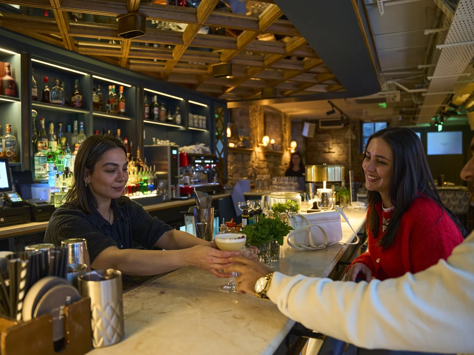 Bar staff member passing a cocktail across a marble bar counter to smiling customers