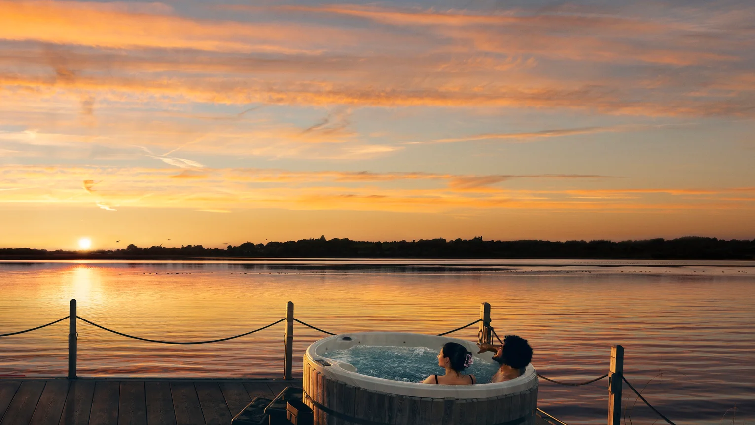 A couple of people in a hot tub on a dock with a sunset