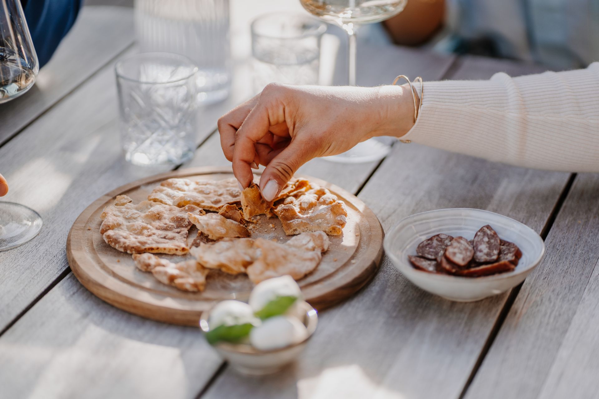 Frau nimmt Stück Südtiroler Schüttelbrot g.g.A. von Holzbrett mit Kaminwurzen, Mozzarella und Gläsern.