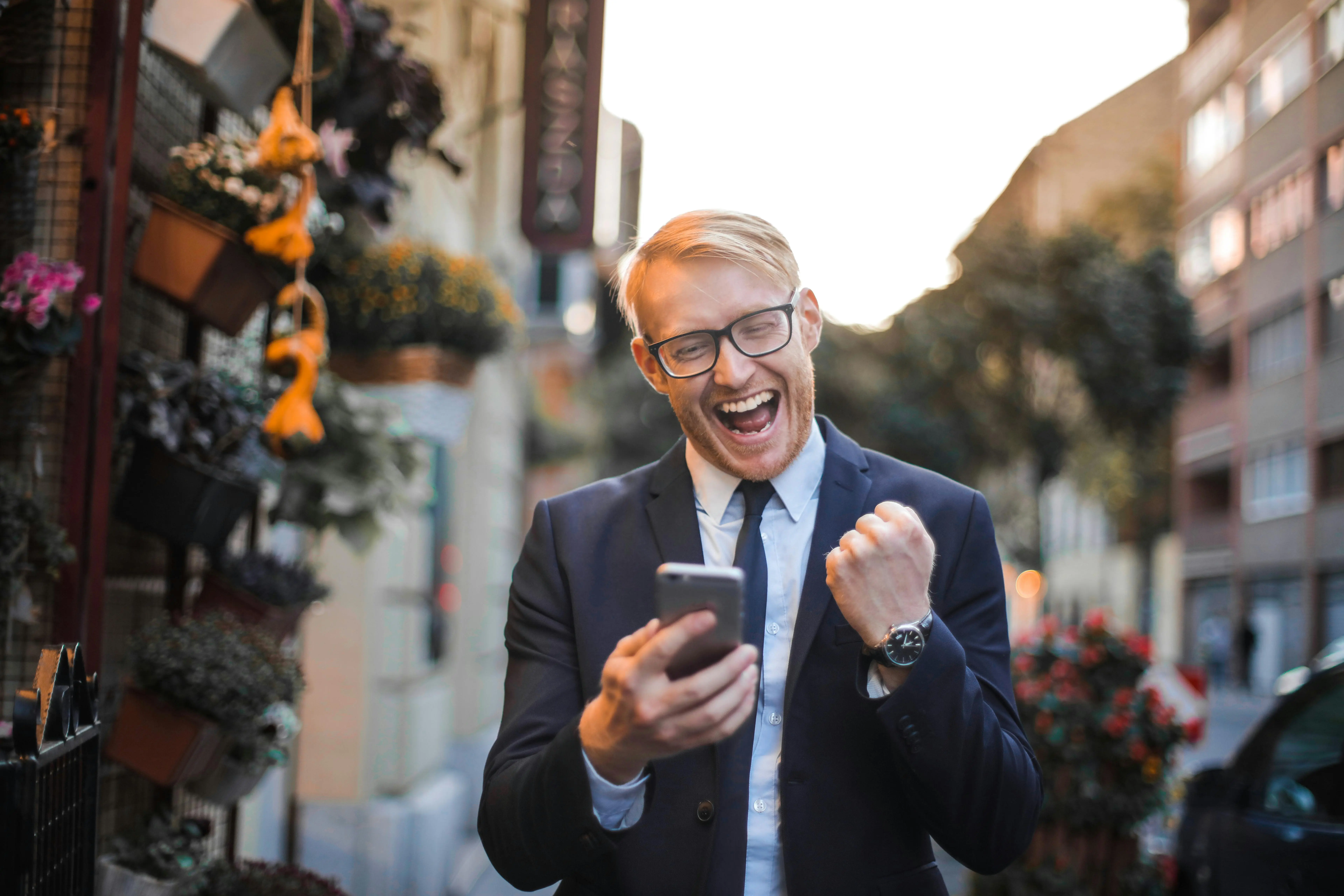 Homme en costume souriant et levant le poing en signe de victoire en regardant son smartphone dans une rue.