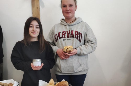 Zwei Schülerinnen halten bäuerlichen Produkten in ihren Händen und im Vordergrund steht eine Käseplatte und Brot. Zwei Schülerinnen halten bäuerlichen Produkten in ihren Händen und im Vordergrund steht eine Käseplatte und Brot.