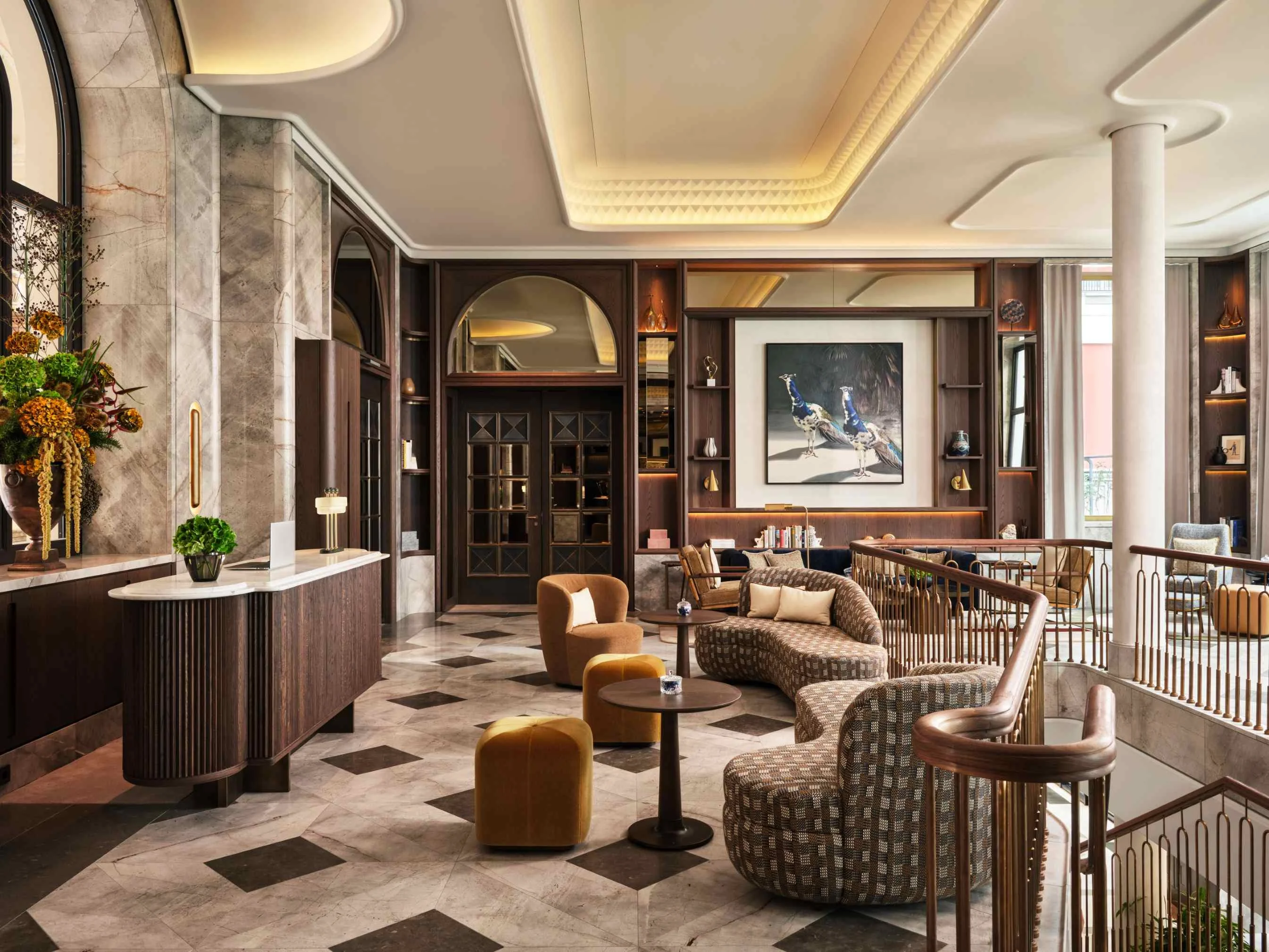 Elegant hotel lobby with marble floor, curved patterned sofa, mustard velvet stools, and a large framed peacock artwork on the wall.