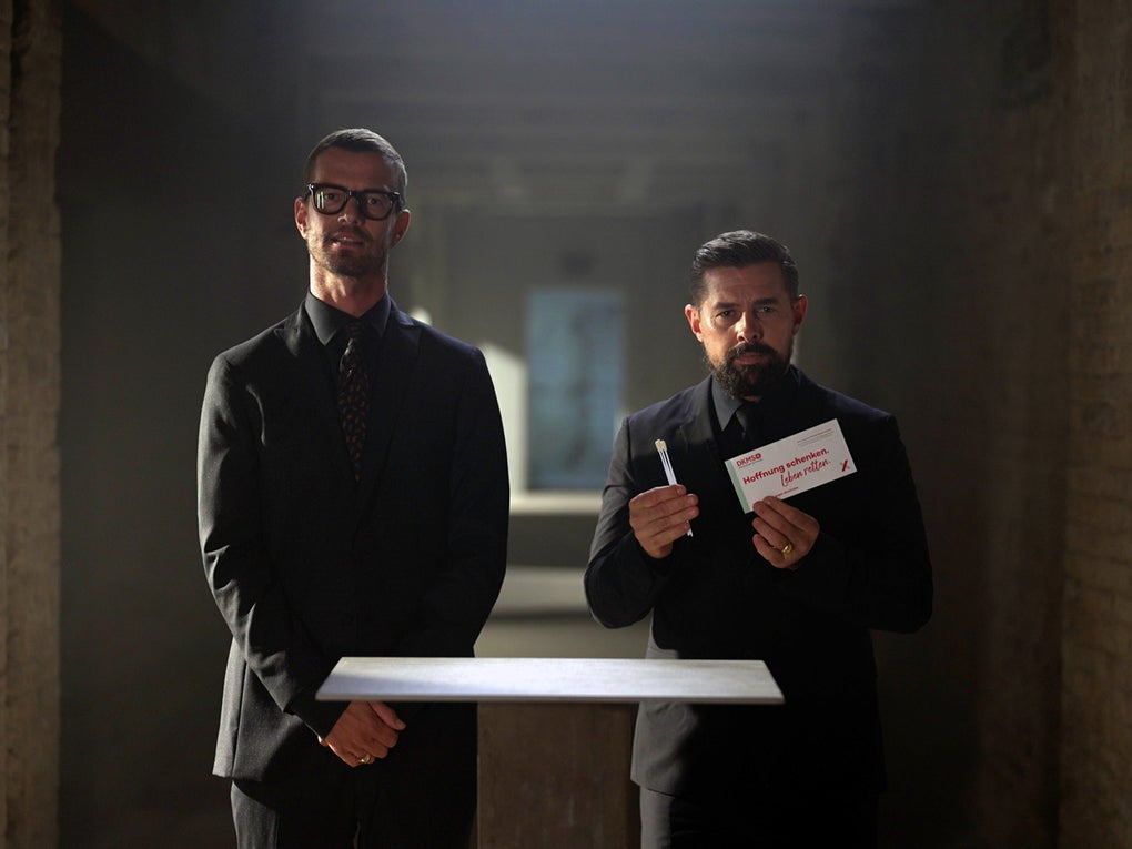 Zwei Männer in dunklen Anzügen stehen nebeneinander in einem schmalen, gemauerten Gang mit Backsteinwänden. Beide haben die Hände vor dem Körper gefaltet. Vor ihnen schwebt ein rechteckiges, weiß leuchtendes Objekt. Der Gang ist nach hinten perspektivisch verlängert und durch Lichtschächte beleuchtet. Zwei Männer in dunklen Anzügen stehen nebeneinander in einem schmalen, gemauerten Gang mit Backsteinwänden. Beide haben die Hände vor dem Körper gefaltet. Vor ihnen schwebt ein rechteckiges, weiß leuchtendes Objekt. Der Gang ist nach hinten perspektivisch verlängert und durch Lichtschächte beleuchtet.