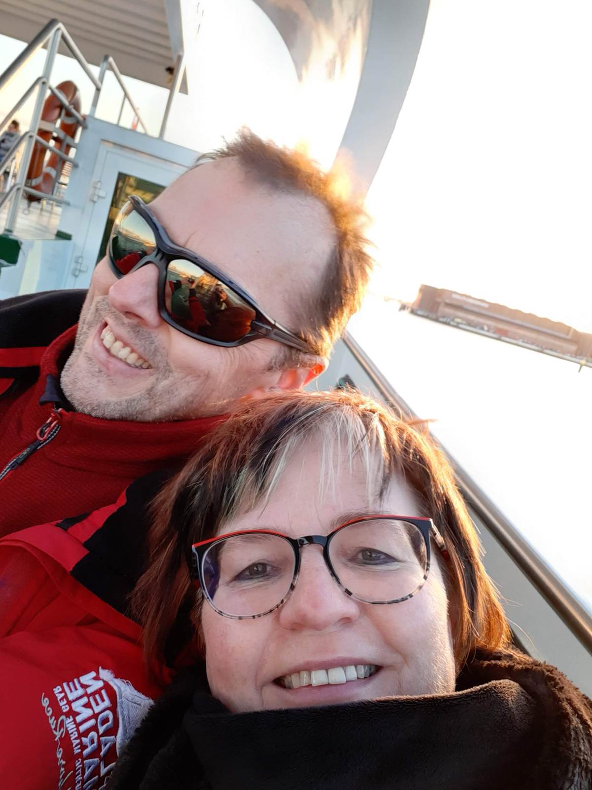 Ein Mann und eine Frau machen ein Selfie an Bord eines Schiffes. Beide tragen Jacken und Brillen, der Mann zusätzlich eine Sonnenbrille. Er trägt eine rote Jacke mit der Aufschrift „Jade Marine“. Im Hintergrund sind Reling, Treppe und Wasser bei Sonnenlicht zu sehen.
Ein Mann und eine Frau machen ein Selfie an Bord eines Schiffes. Beide tragen Jacken und Brillen, der Mann zusätzlich eine Sonnenbrille. Er trägt eine rote Jacke mit der Aufschrift „Jade Marine“. Im Hintergrund sind Reling, Treppe und Wasser bei Sonnenlicht zu sehen.