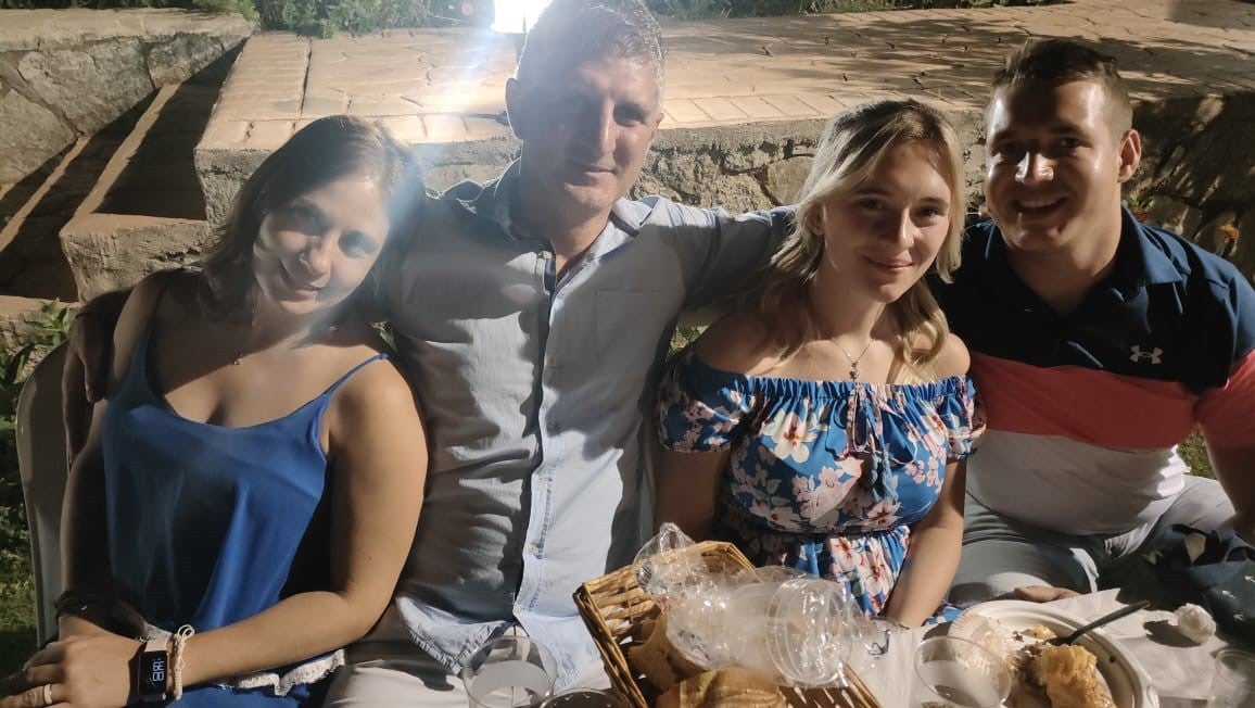 Vier Personen sitzen eng nebeneinander an einem gedeckten Tisch im Freien. Vor ihnen stehen Teller, Gläser und ein Brotkorb. Die Frau links trägt ein blaues Kleid, der Mann neben ihr ein helles Hemd. Rechts daneben sitzt eine Frau in einem geblümten Kleid mit schulterfreiem Ausschnitt, daneben ein Mann im dunkelblauen Poloshirt mit rosa und weißem Streifen. Die Szene ist abends aufgenommen und von künstlichem Licht beleuchtet. Vier Personen sitzen eng nebeneinander an einem gedeckten Tisch im Freien. Vor ihnen stehen Teller, Gläser und ein Brotkorb. Die Frau links trägt ein blaues Kleid, der Mann neben ihr ein helles Hemd. Rechts daneben sitzt eine Frau in einem geblümten Kleid mit schulterfreiem Ausschnitt, daneben ein Mann im dunkelblauen Poloshirt mit rosa und weißem Streifen. Die Szene ist abends aufgenommen und von künstlichem Licht beleuchtet.