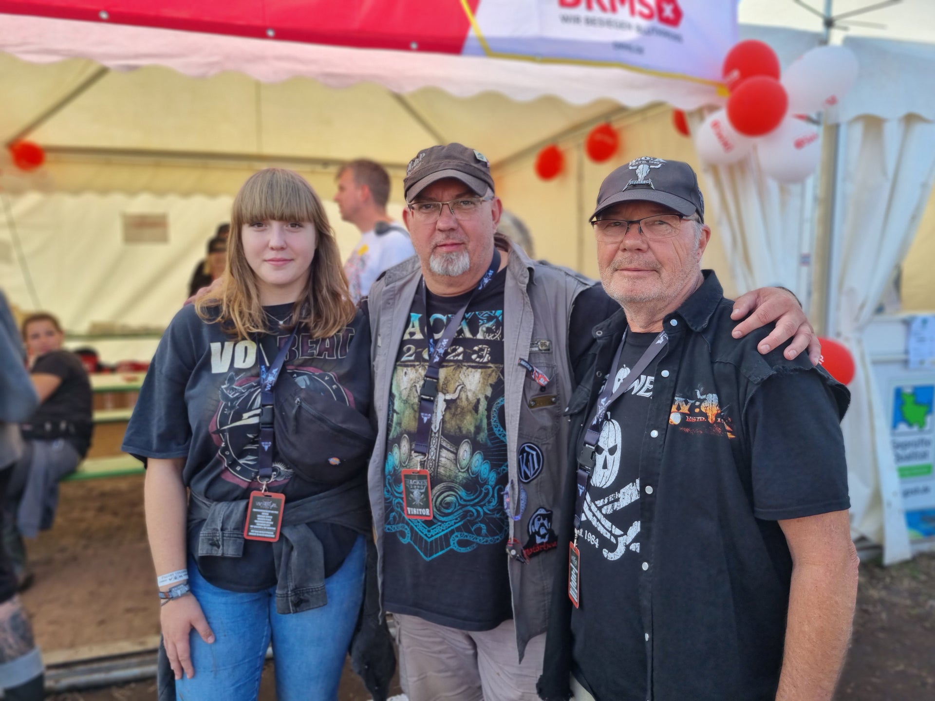 Drei Personen stehen vor einem weißen Zelt mit DKMS-Banner und roten Luftballons. Sie tragen dunkle Kleidung mit Festival- oder Bandmotiven und Festivalpässe um den Hals. Eine junge Frau steht links, zwei ältere Männer stehen rechts, einer mit Arm um die Schulter des anderen. Im Hintergrund sind weitere Personen an Tischen zu sehen. Drei Personen stehen vor einem weißen Zelt mit DKMS-Banner und roten Luftballons. Sie tragen dunkle Kleidung mit Festival- oder Bandmotiven und Festivalpässe um den Hals. Eine junge Frau steht links, zwei ältere Männer stehen rechts, einer mit Arm um die Schulter des anderen. Im Hintergrund sind weitere Personen an Tischen zu sehen.
