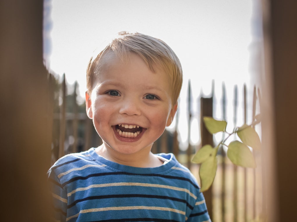 Ein kleiner Junge mit blondem Haar und blau-gestreiftem Shirt steht draußen vor einem Holzzaun. Er lacht in die Kamera. Im Vordergrund sind unscharfe Holzbalken zu sehen, durch die hindurch das Foto aufgenommen wurde. Im Hintergrund sind Zaunspitzen und ein Zweig mit Blättern sichtbar.
Ein kleiner Junge mit blondem Haar und blau-gestreiftem Shirt steht draußen vor einem Holzzaun. Er lacht in die Kamera. Im Vordergrund sind unscharfe Holzbalken zu sehen, durch die hindurch das Foto aufgenommen wurde. Im Hintergrund sind Zaunspitzen und ein Zweig mit Blättern sichtbar.