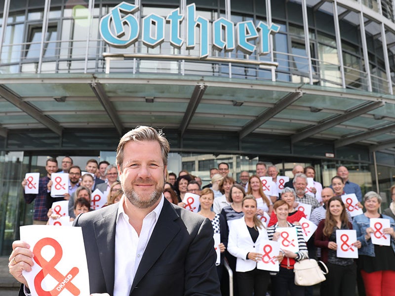 Eine Gruppe von Menschen steht vor einem Gebäude der Gothaer. Alle halten weiße Zettel mit dem Logo der DKMS hoch. Eine Gruppe von Menschen steht vor einem Gebäude der Gothaer. Alle halten weiße Zettel mit dem Logo der DKMS hoch.
