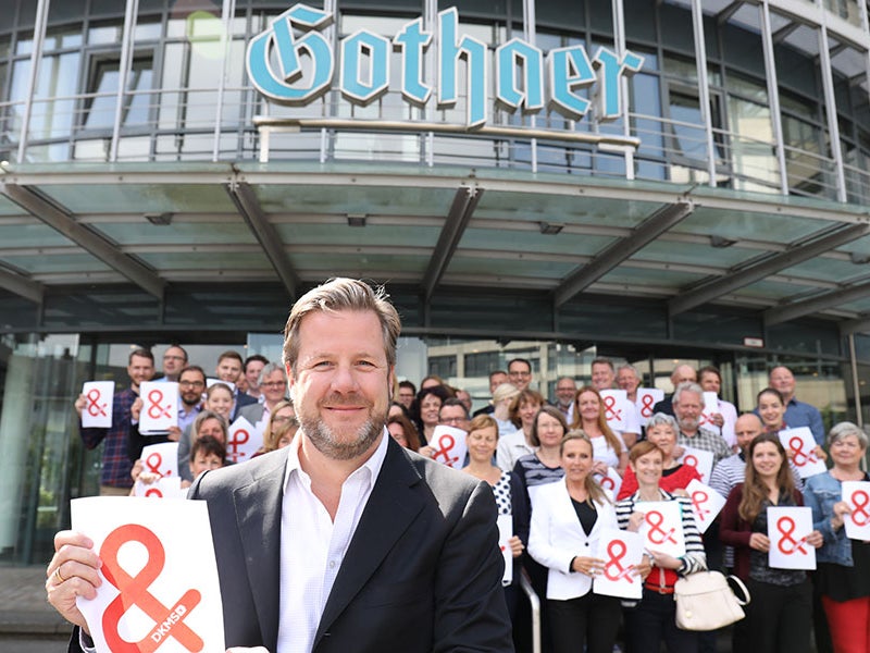 Eine Gruppe von Menschen steht vor einem Gebäude der Gothaer. Alle halten weiße Zettel mit dem Logo der DKMS hoch.  Eine Gruppe von Menschen steht vor einem Gebäude der Gothaer. Alle halten weiße Zettel mit dem Logo der DKMS hoch.